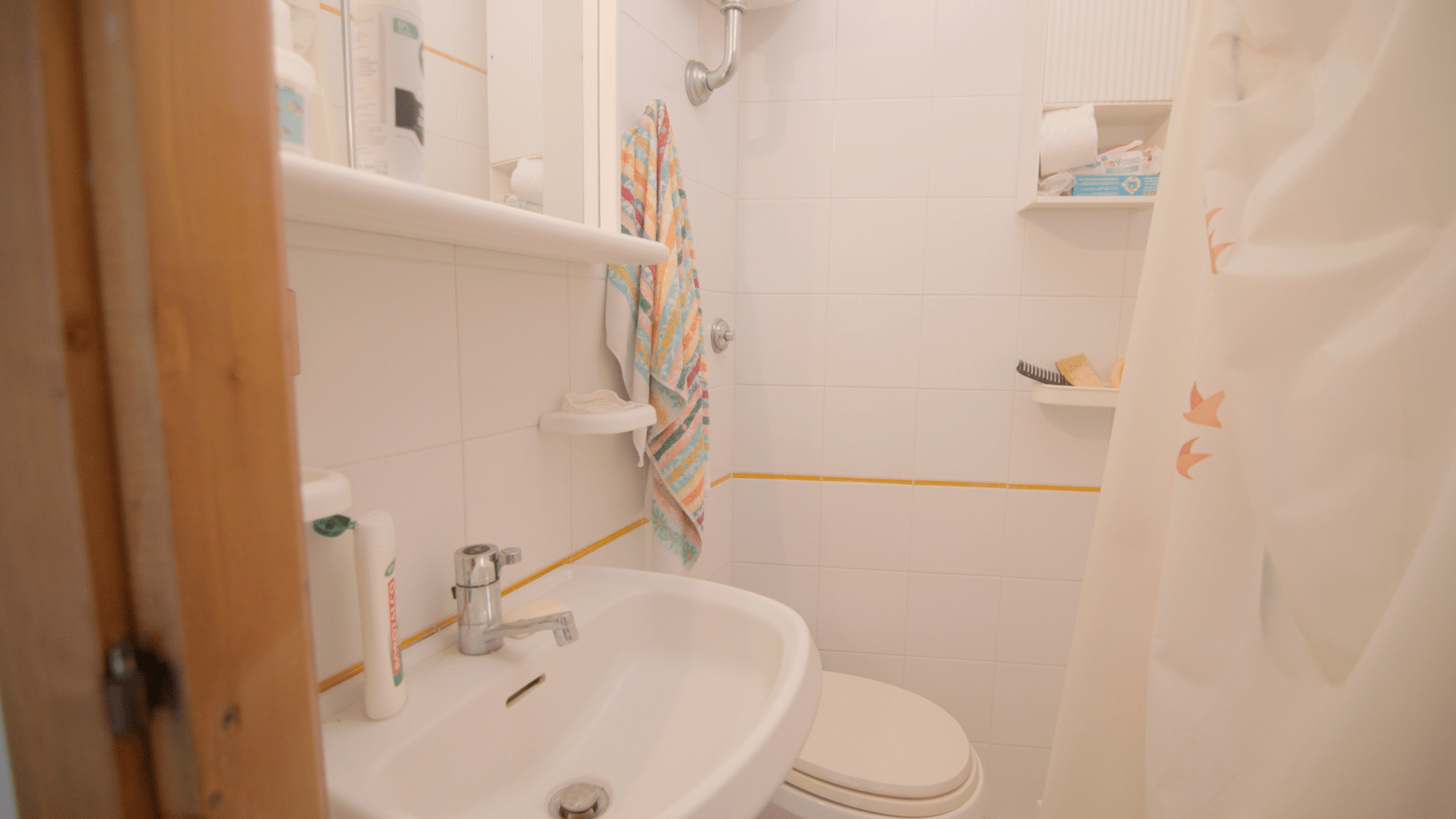Small tiled bathroom with a sink, toilet, shower curtain, and a colorful towel hanging on the wall.