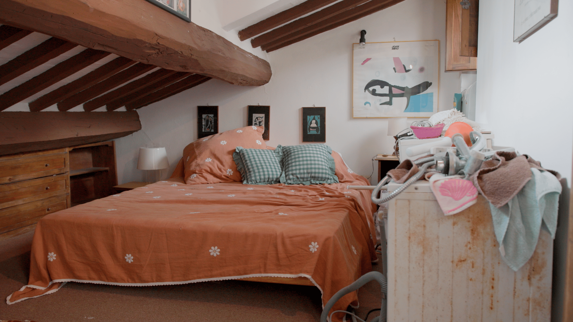 Rustic attic bedroom with orange bed, wooden beams, drawers, art, and laundry on the right.