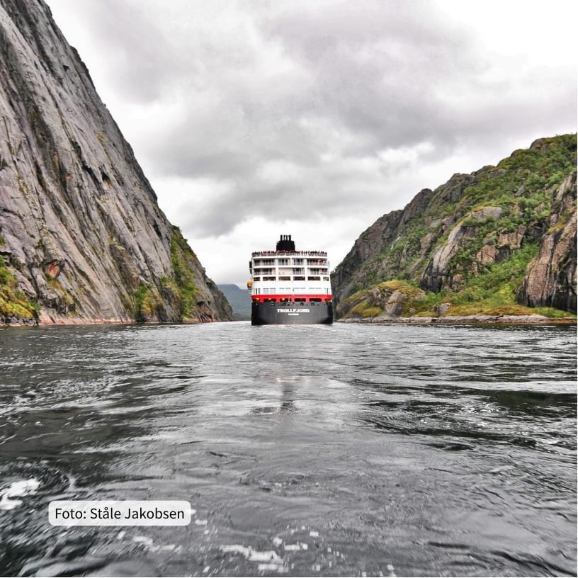 The large cruise ship Trollfjord navigates a dramatic narrow fjord framed by steep rocky mountains.