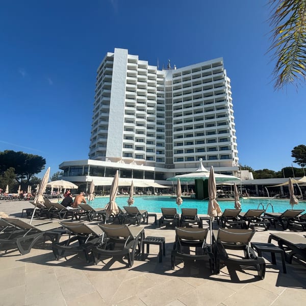 A grand white hotel overlooks a resort pool with many lounge chairs and umbrellas under a clear blue sky.
