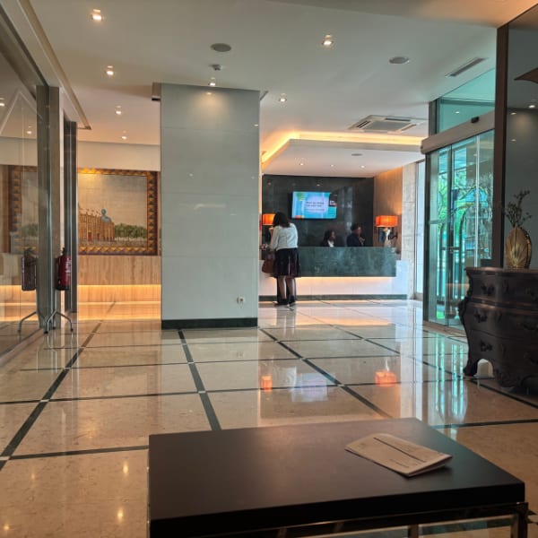 A bright hotel lobby with a woman at the reception desk, staff, and a large tiled floor.