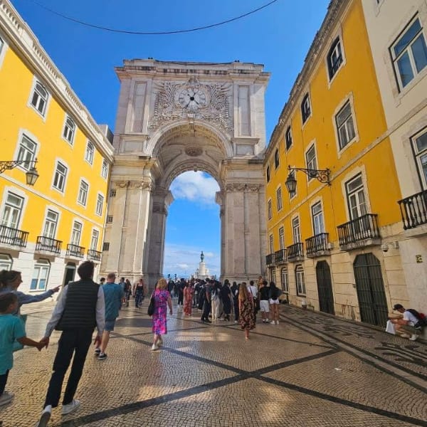People walk towards Lisbon's grand Rua Augusta Arch, framed by bright yellow buildings under a blue sky.