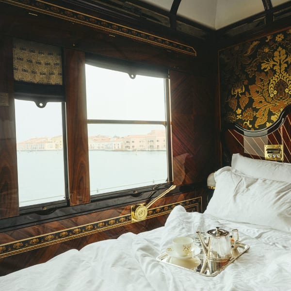 Luxurious train cabin bedroom with a made bed, silver tea set, and a window view of a canal city.