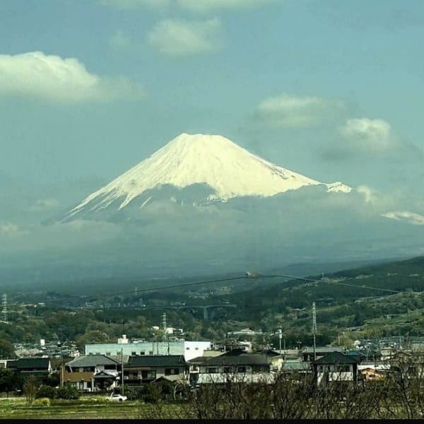 Snow-capped Mount Fuji towering over a residential area and green hills under a cloudy sky.