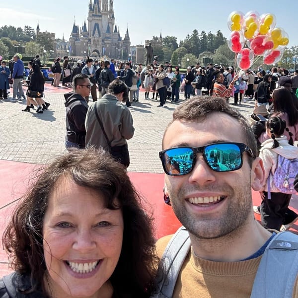 Selfie of a woman and smiling man in sunglasses, castle and crowd behind them.