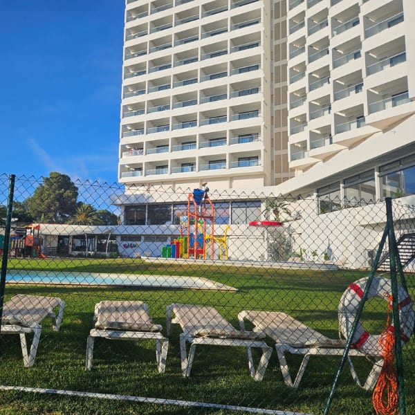 A resort scene with a tall white hotel, kids' play area, pool, and lounge chairs viewed through a chain-link fence.