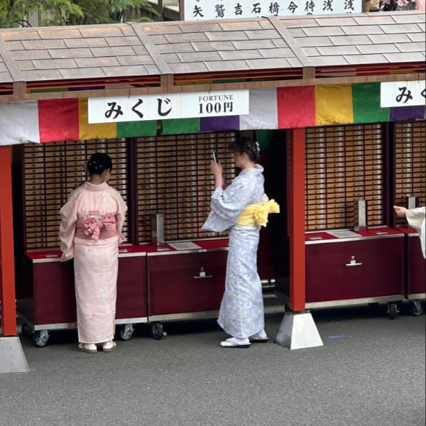 Two women in kimonos at a shrine's fortune slip stand, one takes a photo.
