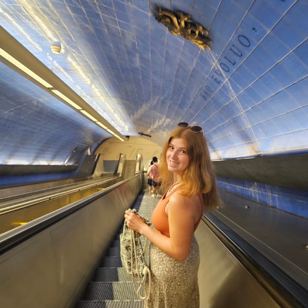 A smiling young woman in a floral skirt and orange top poses on a long escalator in a blue-tiled tunnel.