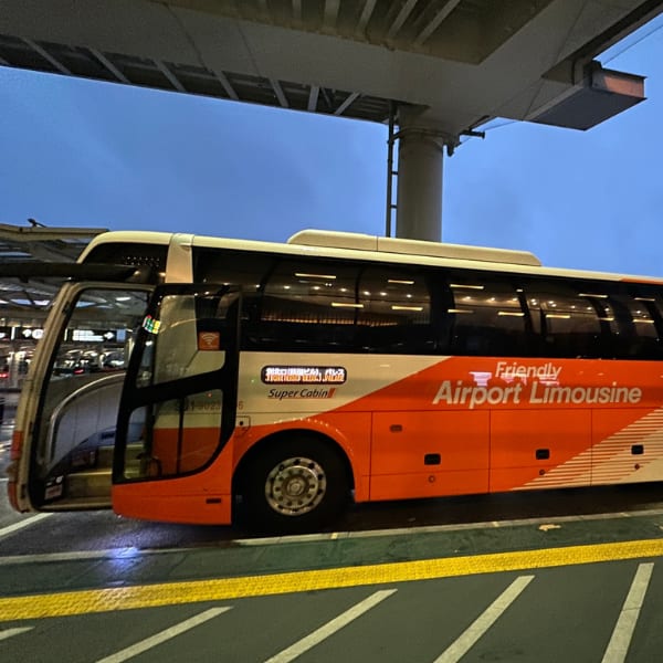 Orange and white Friendly Airport Limousine bus with open door, parked under an elevated structure.