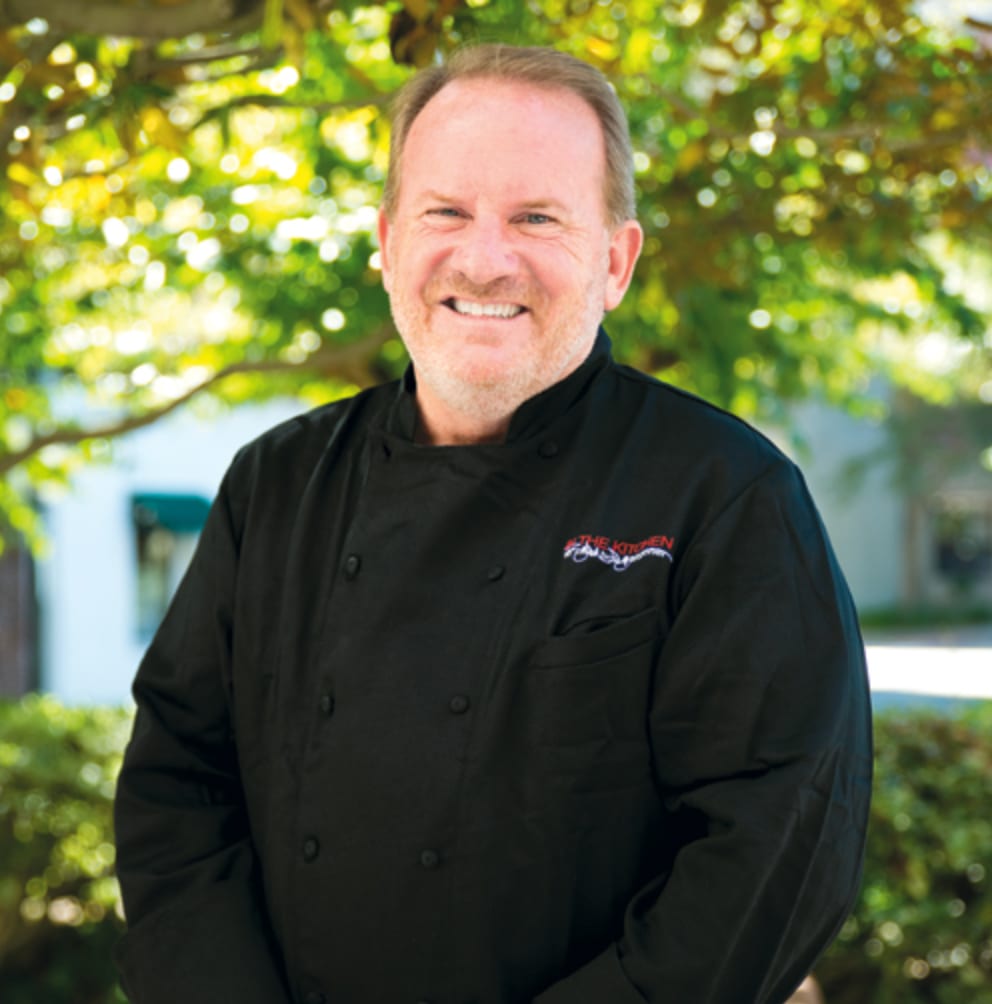 Smiling chef in black jacket with "THE KITCHEN" logo.