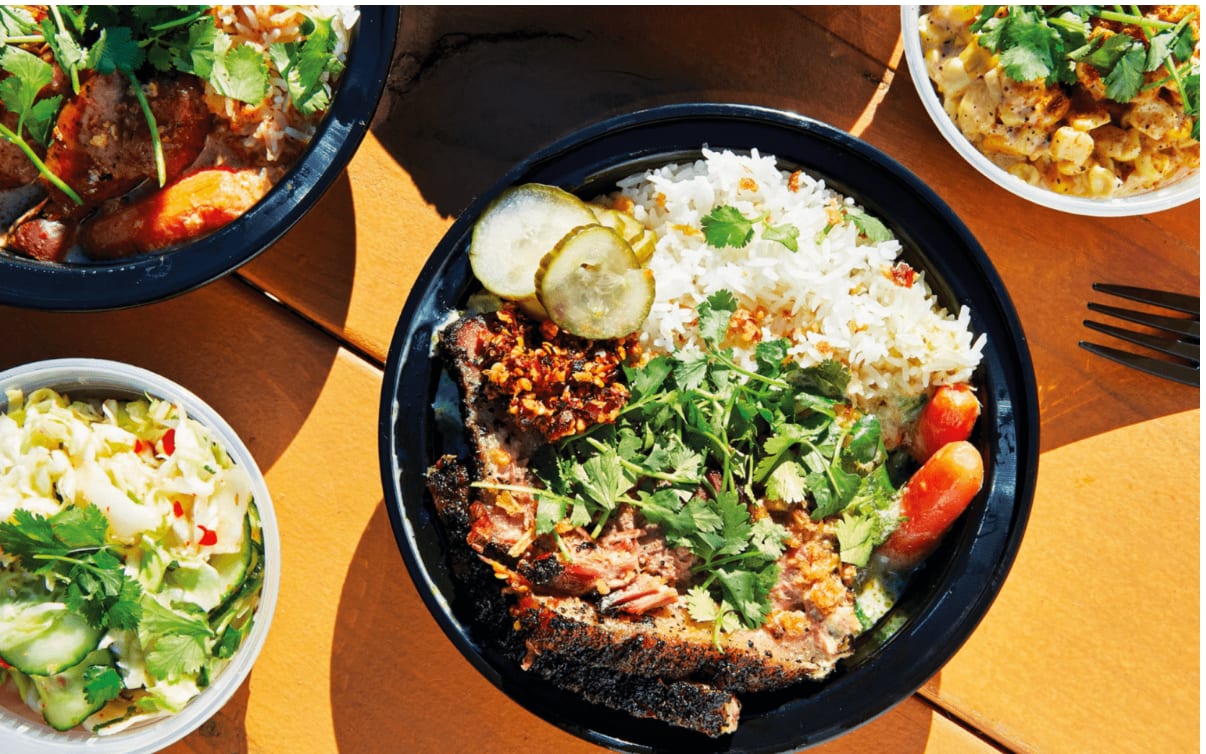 Top-down view of multiple food bowls on a wooden table. Main bowl has rice, seared protein, and sides.