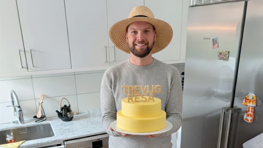 Smiling man in a straw hat holding a yellow cake with "TREVLIG RESA" text on top and on his sweater.