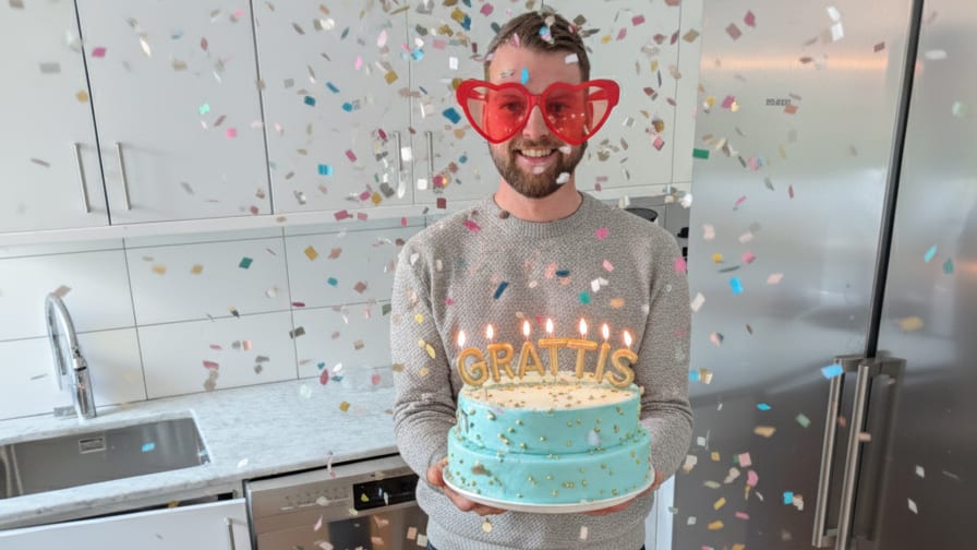 Man in heart glasses holds a birthday cake with "GRATTIS" candles as confetti falls in a kitchen.