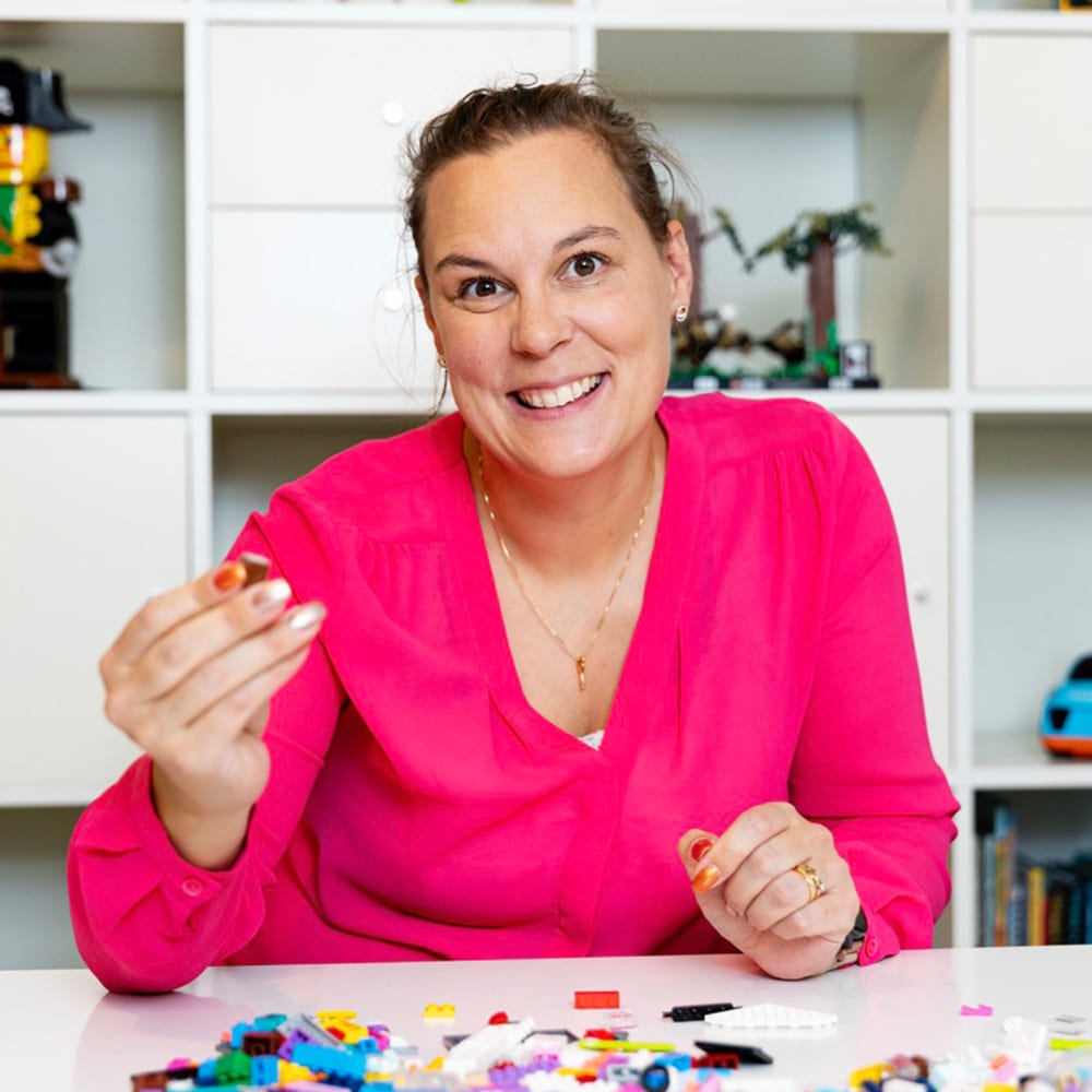 Smiling woman holding a Lego piece, with colorful bricks on a table and models behind her.
