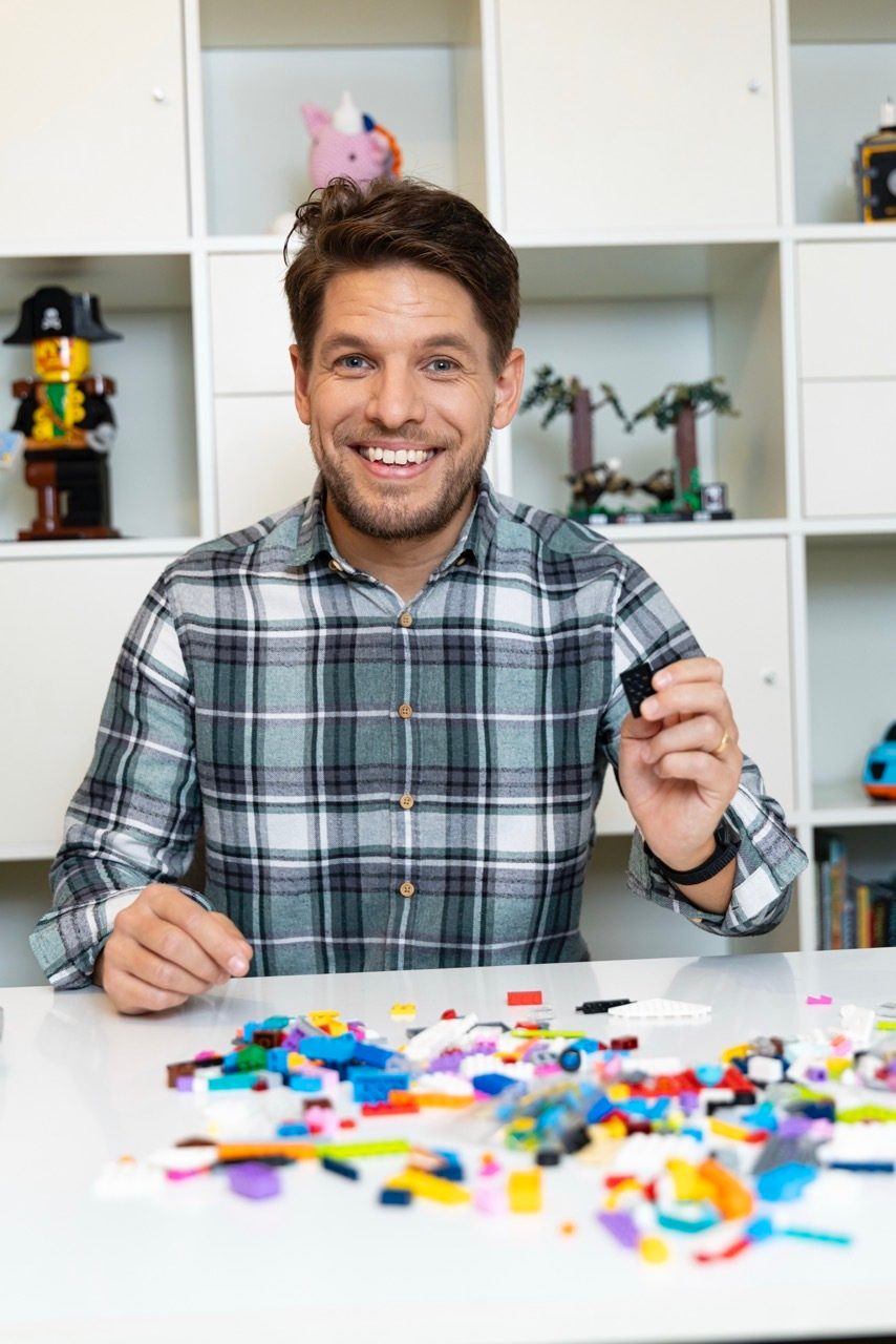 Smiling man in plaid shirt holding a Lego piece, surrounded by colorful Lego bricks.