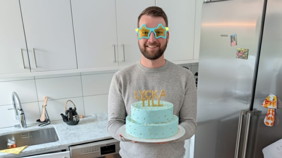 Man with star glasses holds blue cake inscribed "LYCKA TILL" in kitchen.
