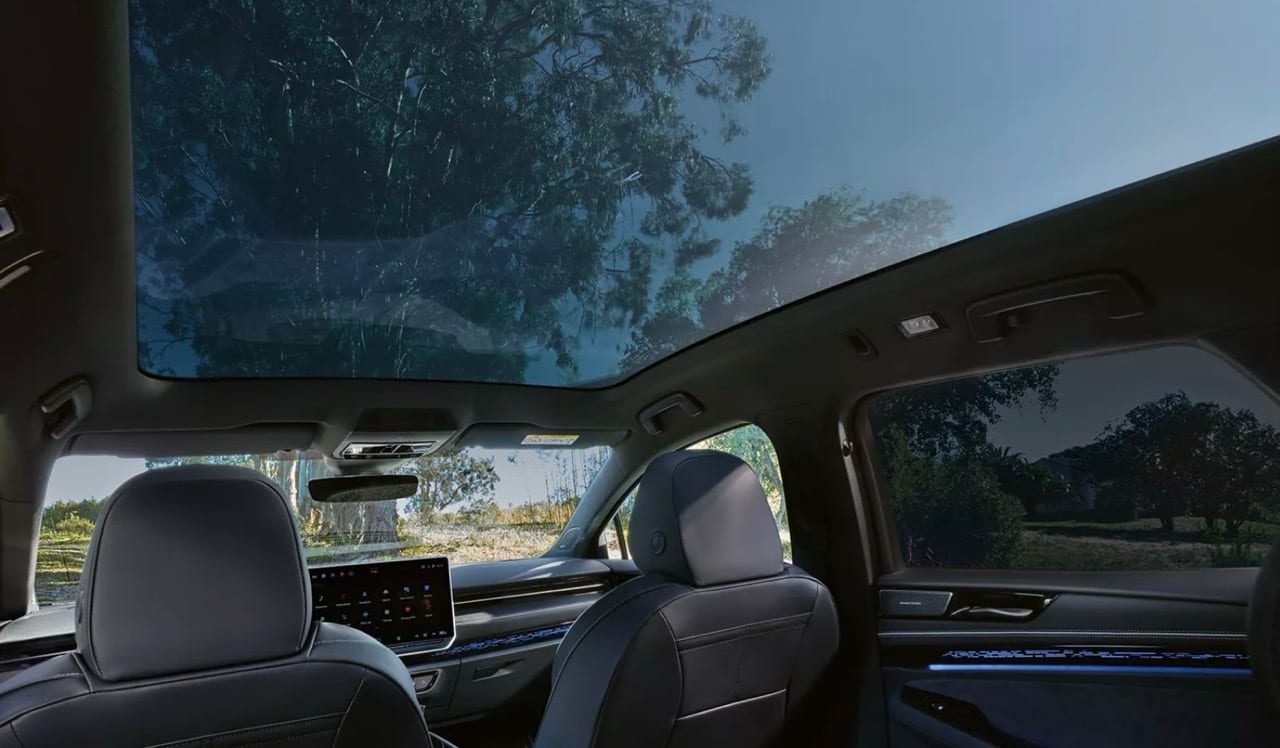 Car interior with panoramic glass roof, showing trees and sky outside, with seats and dashboard.