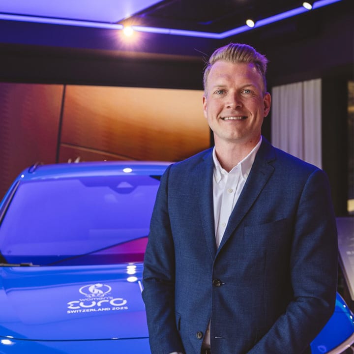 Man in blue suit smiles by blue car with Women's EURO 2025 logo.