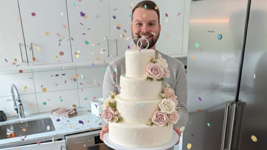 Smiling man holding a four-tier wedding cake with flowers and confetti falling around him.