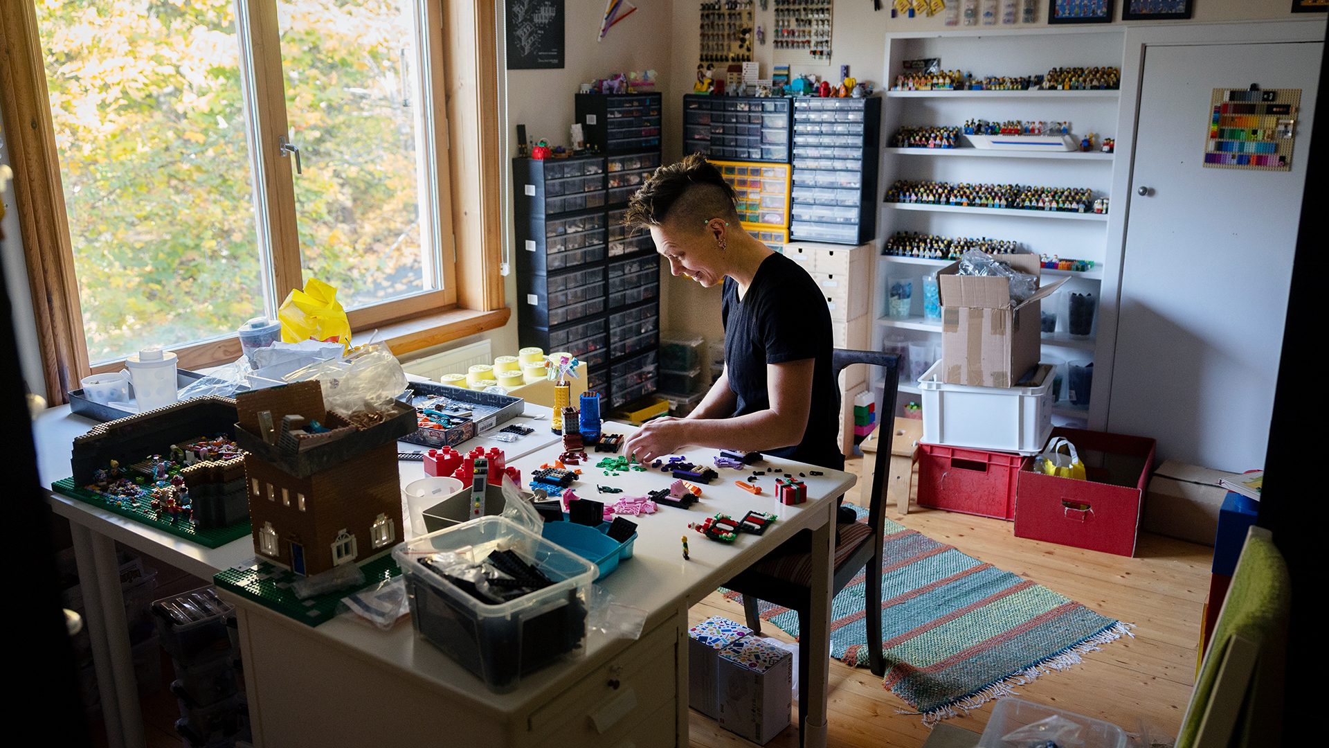 A person builds Lego models at a desk in a room organized with extensive Lego storage and minifigures.