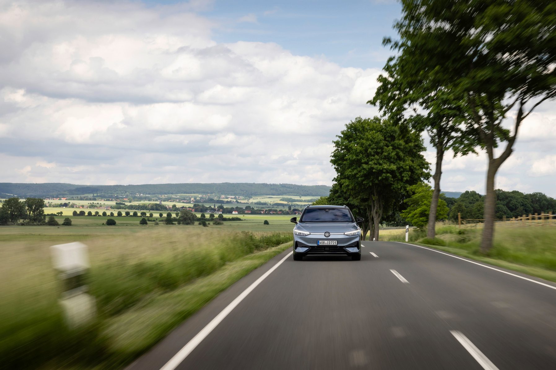 Grey car on a scenic road, motion blur, green fields, trees, and distant hills.