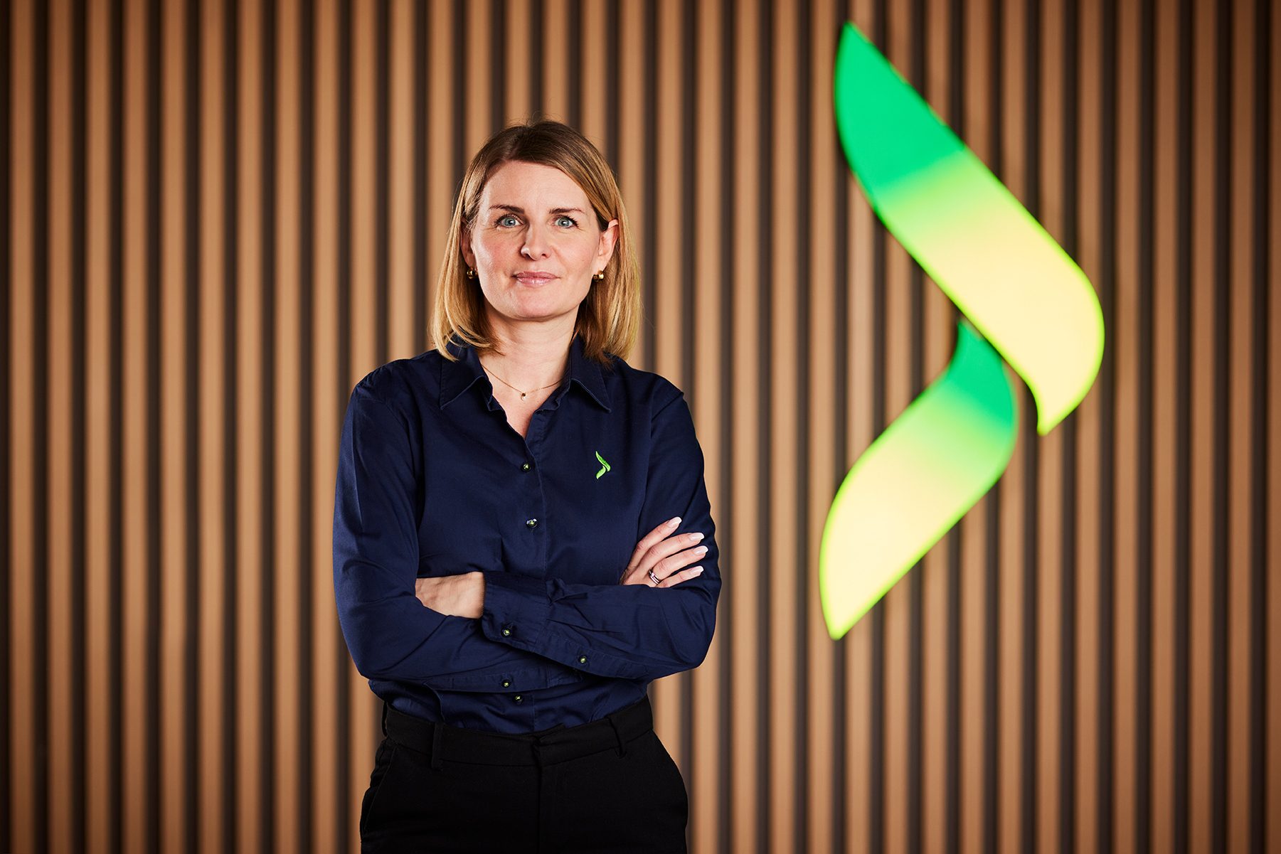 Woman in blue shirt with crossed arms, standing before a striped wall with a prominent green and yellow logo.