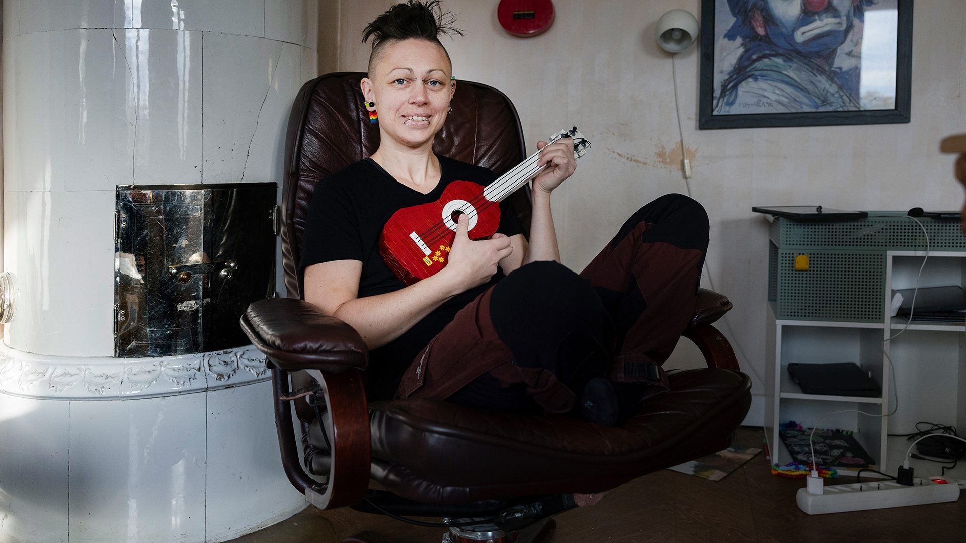 Smiling person with a mohawk and piercings holds a red ukulele while sitting in a chair.