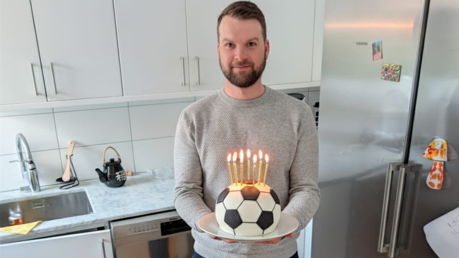 A man holds a soccer ball cake with lit candles in a kitchen.
