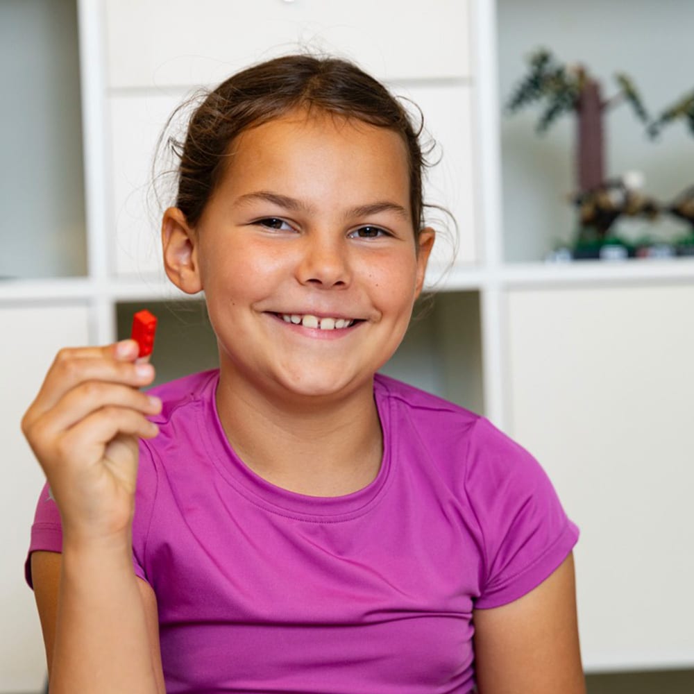 Smiling girl in purple shirt holding a red Lego brick.