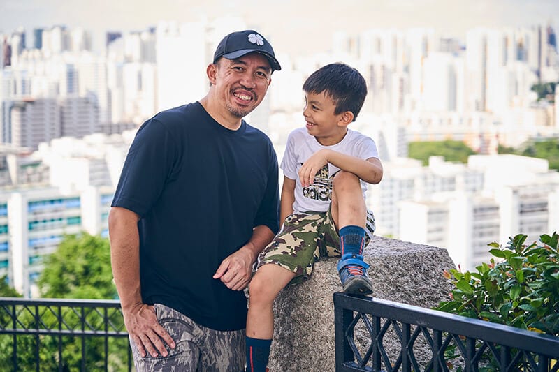 Man smiles at viewer, boy smiles at man, city background.