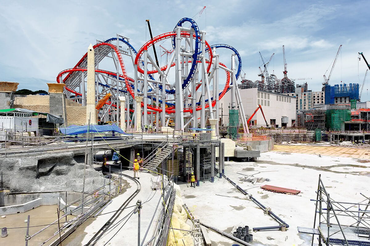 Construction of a theme park with a prominent red and blue rollercoaster and cranes.