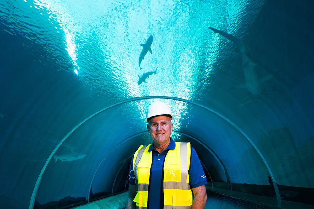Man in hard hat and vest smiles in an underwater tunnel with sharks above.