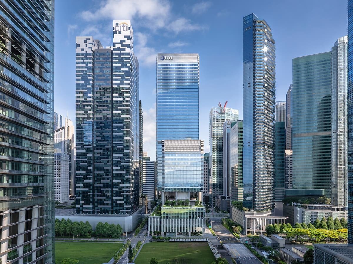 Modern cityscape featuring tall glass skyscrapers, green parks, and urban roads under a blue sky.