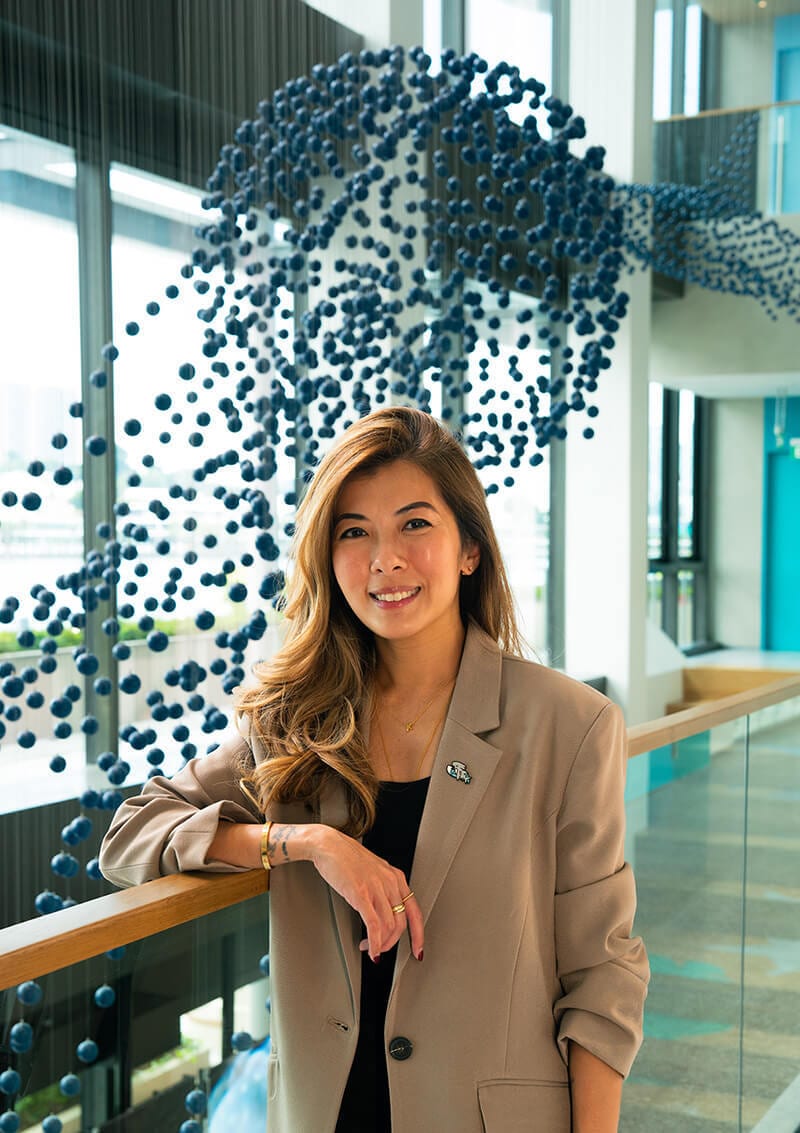 Smiling woman in a beige blazer leans on a railing, blue sphere art in background.