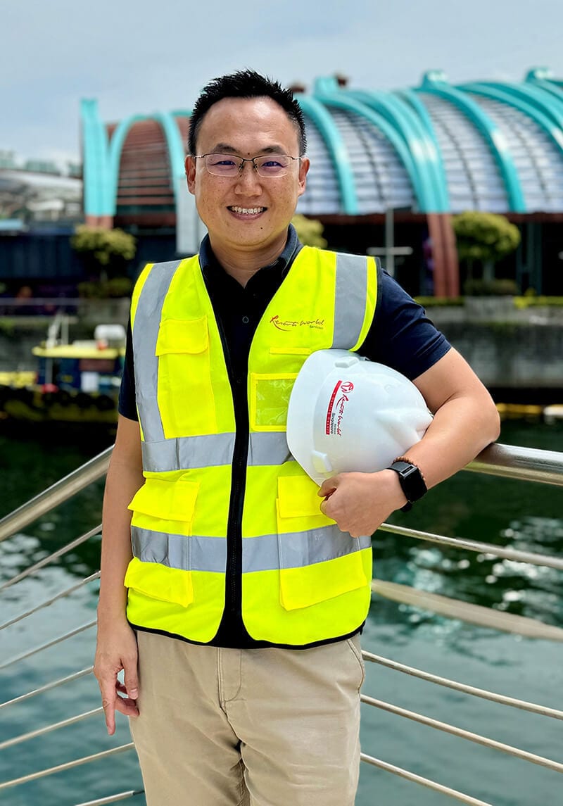 Smiling man in Resorts World safety vest holding hard hat by water.