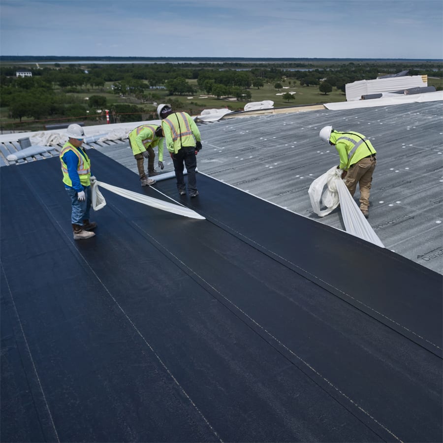 Four construction workers installing black roofing material on a large flat roof.