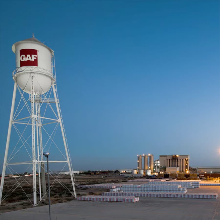 GAF logo on white water tower at dusk with GAF building lit up in distance.