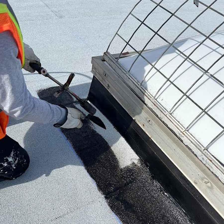 Worker torching black roofing material around a skylight on a white roof.