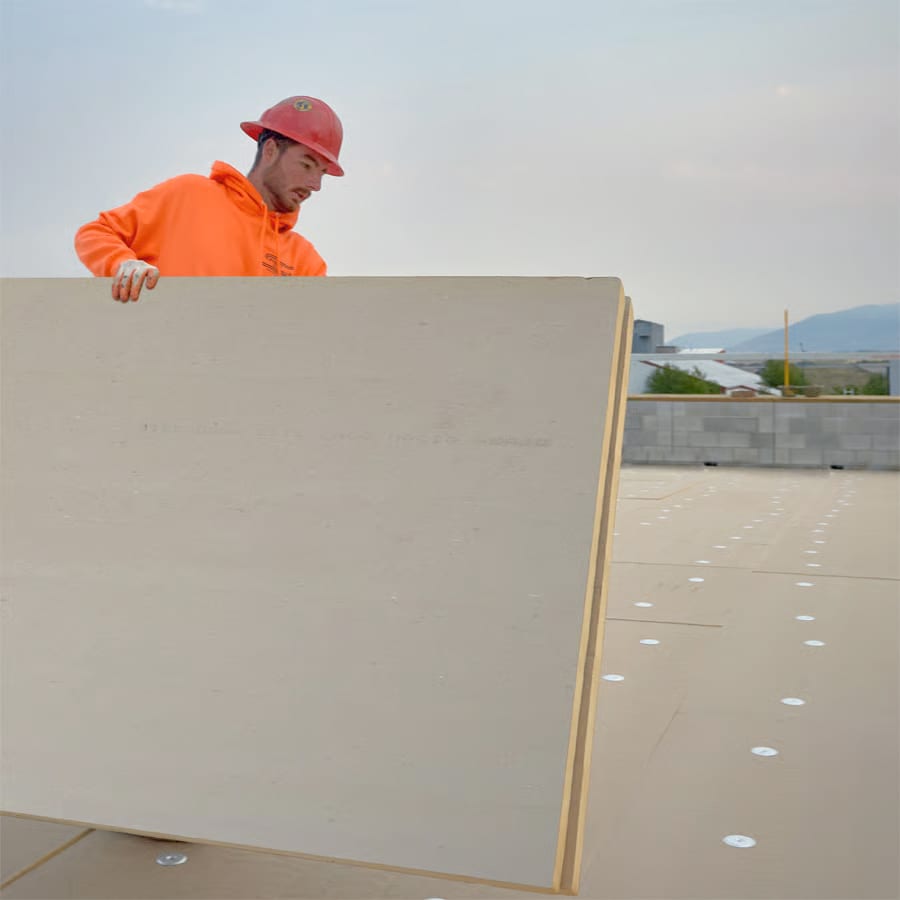 Roofer in orange sweatshirt and red hard hat carrying coverboard on a roof.