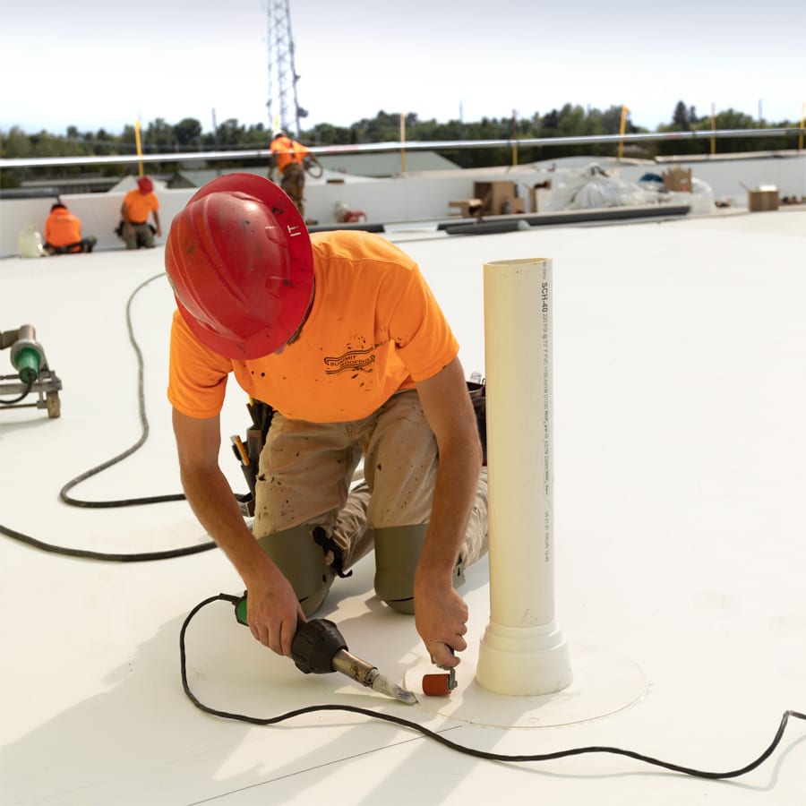 A roofer from Summit Roofing kneels, using a heat welding tool and roller to seal a PVC pipe on a commercial roof.