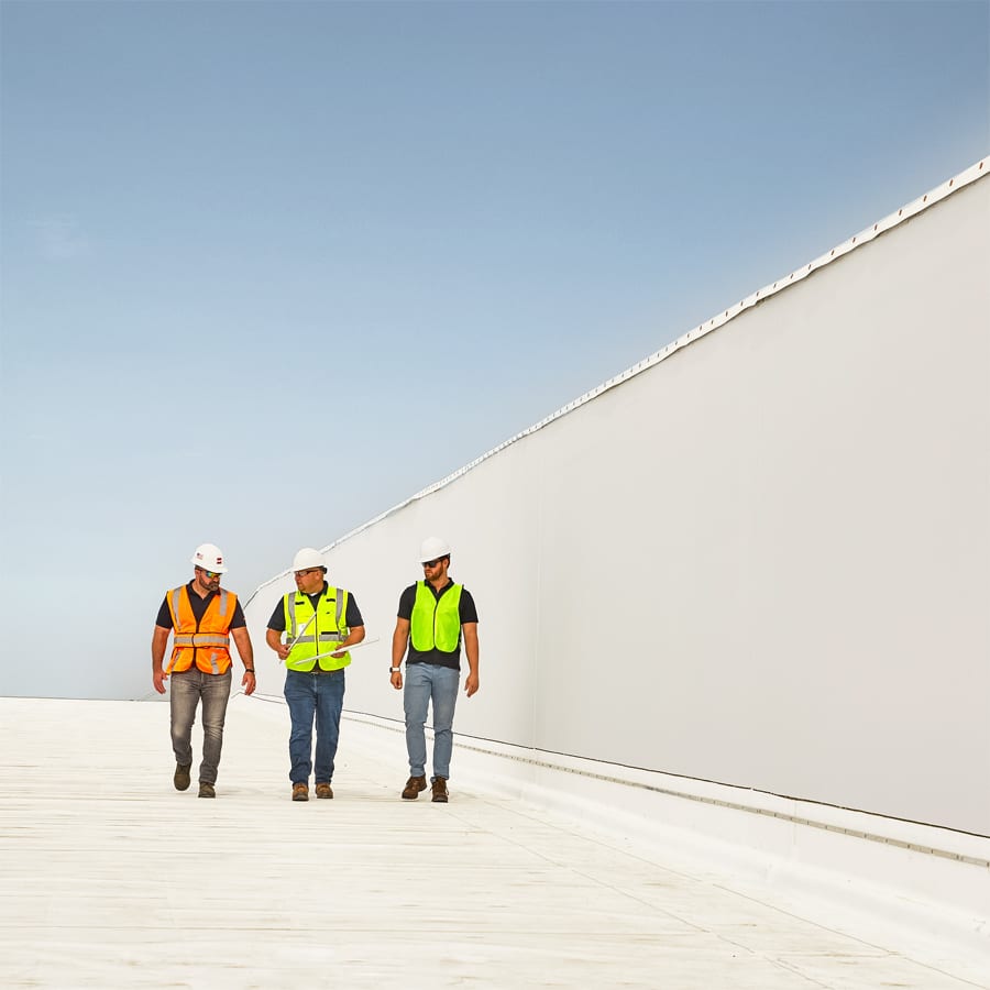 Three workers in safety gear walk on a white building roof.