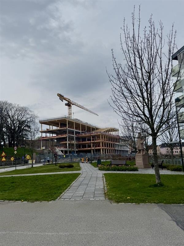 Large building under construction with crane, green park with statue and bare trees.