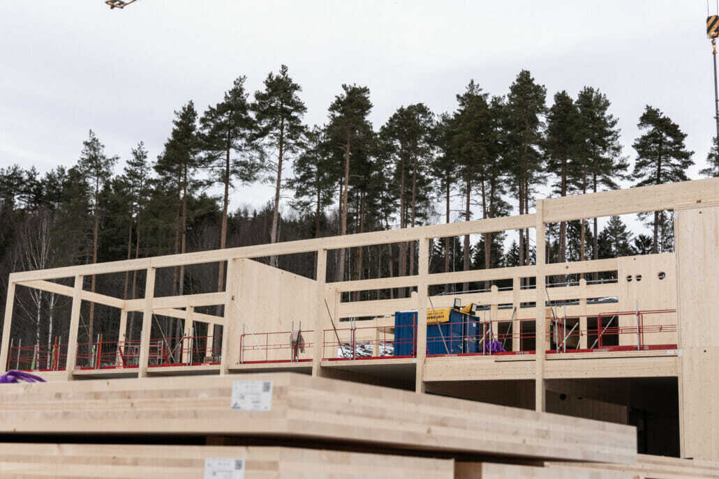 Timber frame building under construction, with stacked wood and a forest in the background.