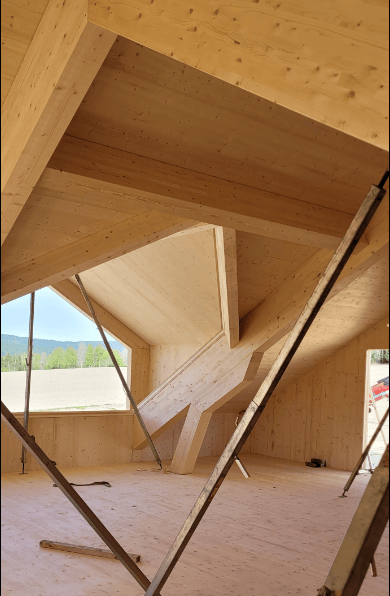 Interior wooden building under construction, showing large beams, temporary supports, and a window.