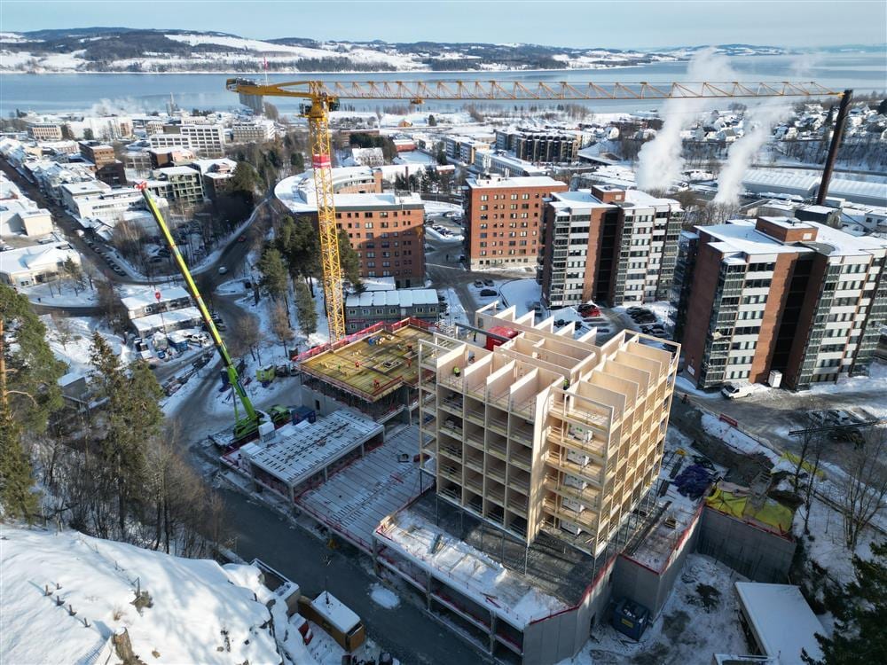 Construction of a large wooden building in a snowy city by a lake, with two cranes.