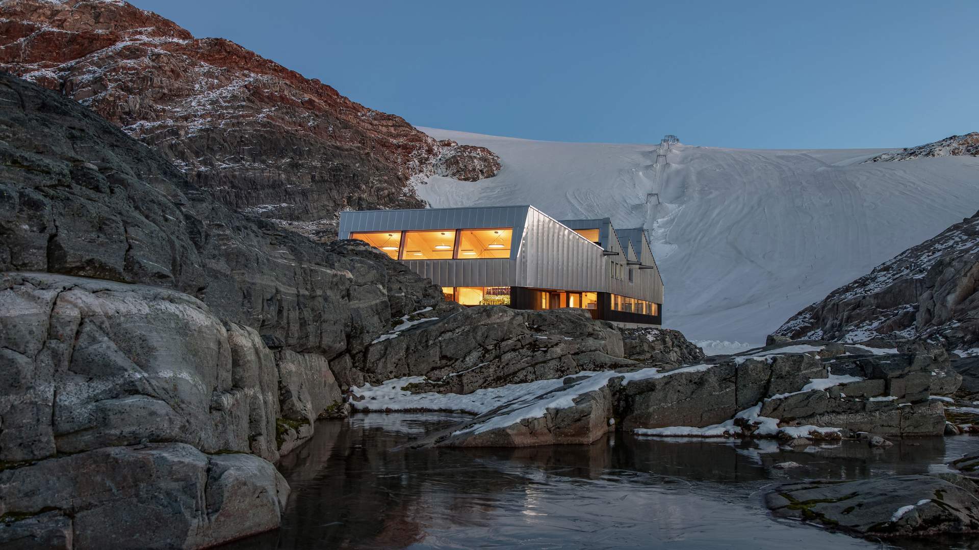 Modern hut with warm lights in a snowy, rocky mountain landscape by a partially frozen pond.