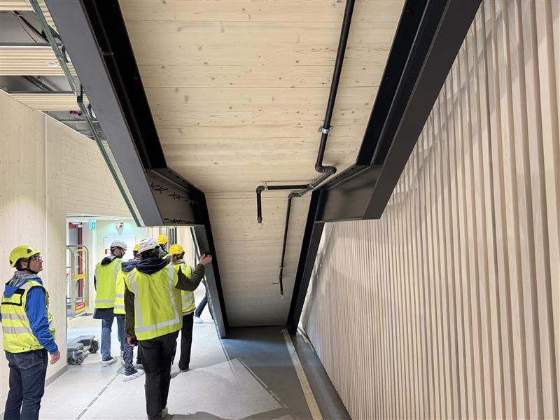 Site workers observe a building with a black metal beam, wooden ceiling, and slatted wall.