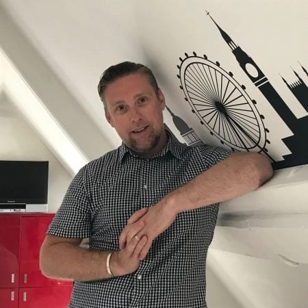 Man in plaid shirt smiling, leaning against a wall with a London skyline decal.