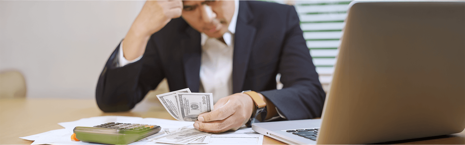 Stressed man in a suit counting money at a desk with a calculator and laptop.