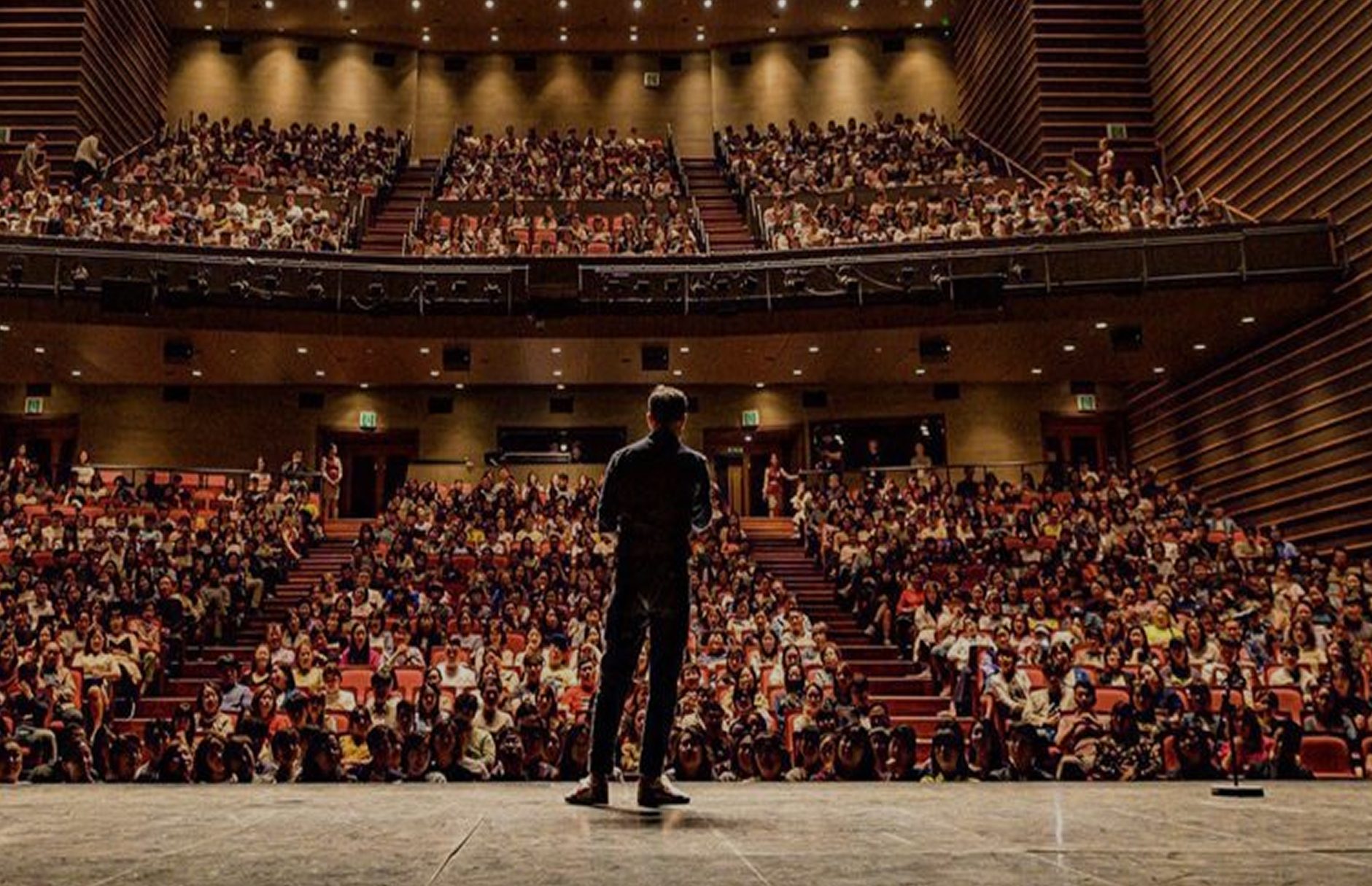 A person stands on stage with their back to the viewer, addressing a large audience in an auditorium.